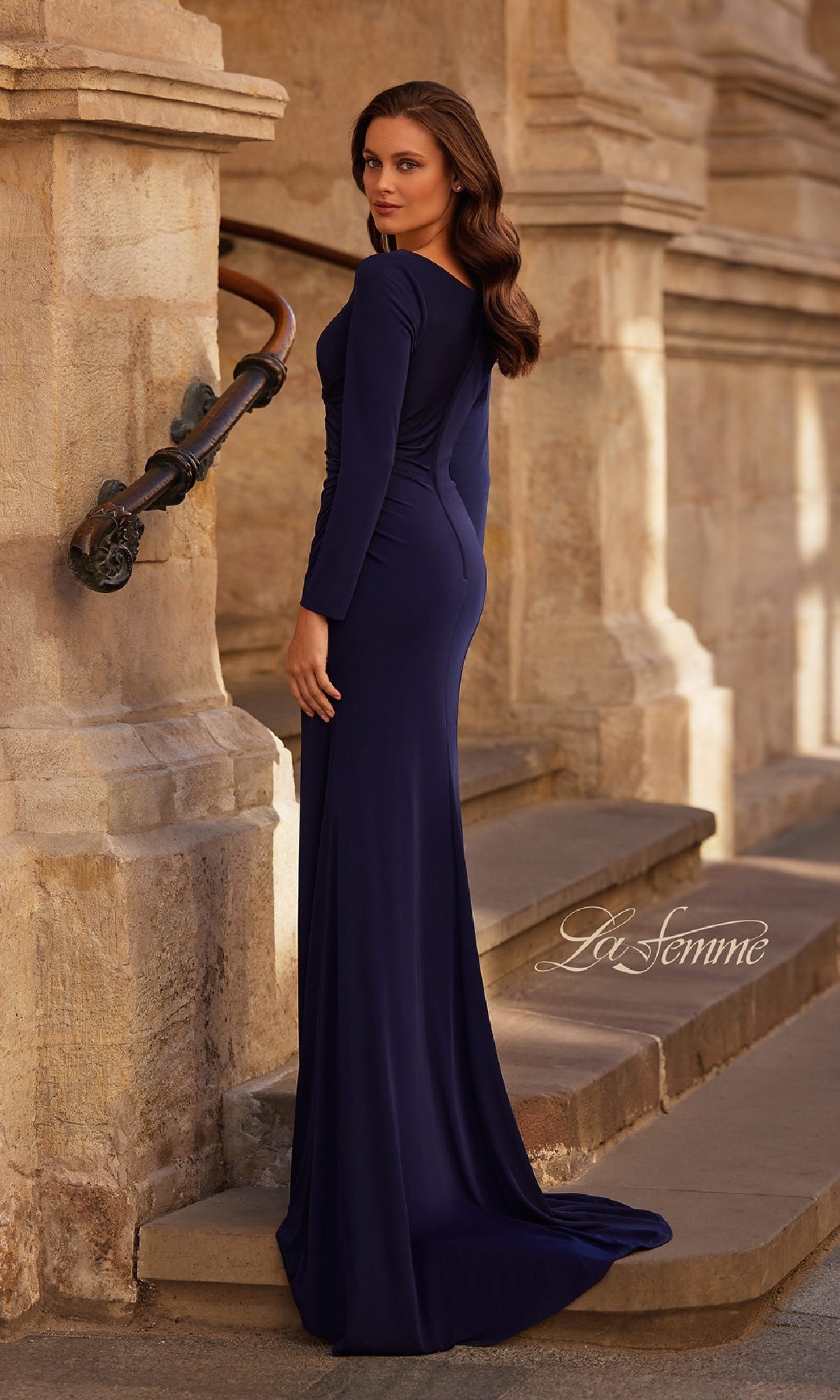 A woman wearing the La Femme 29184 Long Sleeve Long Formal Dress stands on stone steps, glancing over her shoulder with loose waves in her hair.